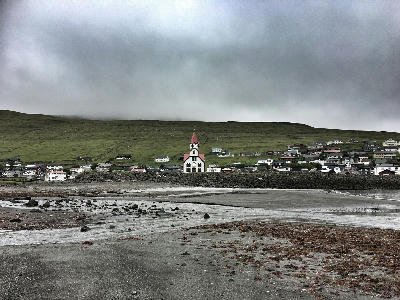 Sørvágur Church with red roof standing behind a stone seawall, viewed across a shallow tidal shoreline with village houses and green hillside on Vágar Island, Faroe Islands.