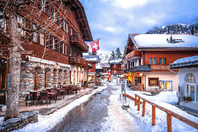 Snow-covered street in Mürren village with wooden chalets, Swiss flags, and outdoor seating along the walkway, Switzerland.