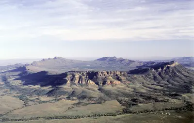Aerial view of Wilpena Pound in the Flinders Ranges, South Australia, showing rugged sandstone escarpments, a broad basin and distant mountain ranges beneath a lightly clouded sky.