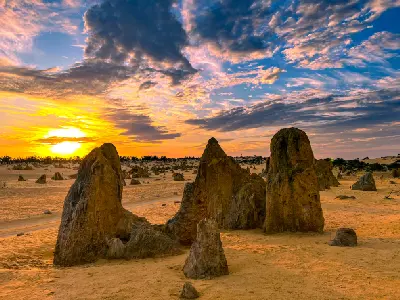 Limestone pinnacles rise from golden desert sand beneath a dramatic sunset sky.