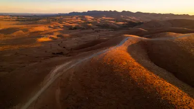 Aerial view of a winding road crossing sunlit ochre hills near Razorback Lookout in the Flinders Ranges, South Australia, at golden hour.