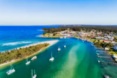 Aerial view over turquoise Currambene Creek with sailing boats and a sandy spit, opening into Jervis Bay beside the waterfront town of Huskisson.