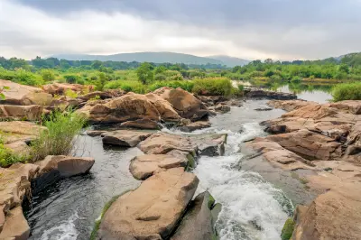 A scenic landscape showing a rushing stream cascading through smooth, light brown rock formations and boulders on the Crocodile River in Kruger National Park. The lush green riverbanks feature reeds and trees under an overcast sky, with the rolling green ridges of the Lebombo Mountains visible along the horizon line.