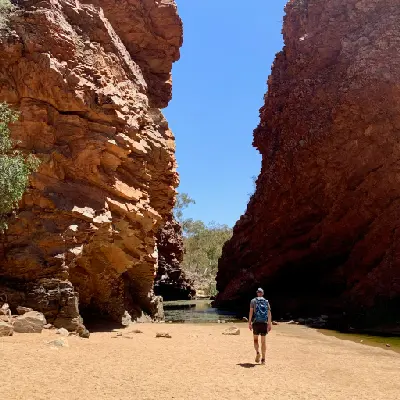 A lone hiker with a backpack is dwarfed by immense, rugged red rock walls rising high on either side at Simpsons Gap, or Rungutjirpa.