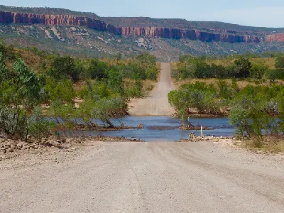 Gibb River Road descends through the shallow Pentecost River with the red escarpments of the Cockburn Range rising beyond.
