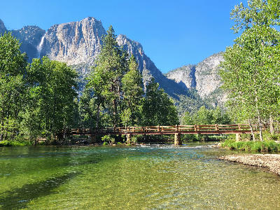 Sentinel Bridge crossing the Merced River with clear green water, trees, and Upper Yosemite Fall in Yosemite National Park.