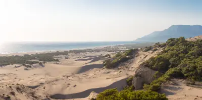Sweeping view across the rolling sand dunes of Patara toward its long Mediterranean shoreline, with pine-covered slopes and mountains rising in the distance.