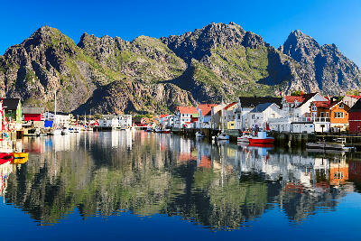 Colourful fishing houses and boats reflected in the harbour at Henningsvær on Austvågøya in the Lofoten Islands, with steep granite peaks rising behind in Nordland, Norway.
