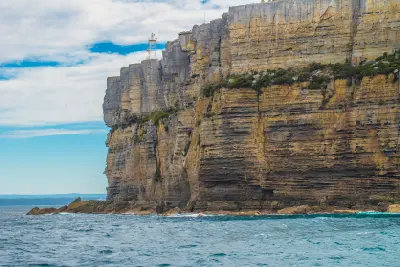Towering layered sandstone cliffs at Point Perpendicular rise above the ocean, with the lighthouse visible on the clifftop beneath a partly cloudy sky.