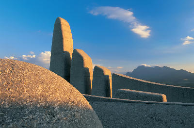 Close-up architectural view of the Afrikaans Language Monument in Paarl, South Africa, showing curved concrete spires lit by warm evening light with mountains and blue sky in the background.