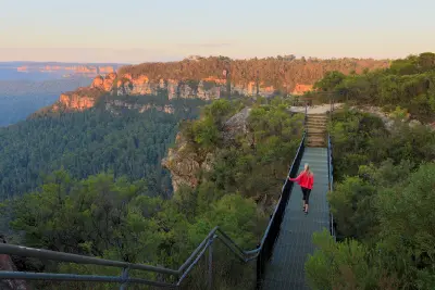Visitor crosses a narrow metal footbridge above the forested Jamison Valley, with sandstone cliffs glowing in warm evening light.