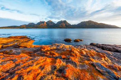 Orange-lichen-covered granite rocks beside calm blue water with The Hazards mountain range glowing in evening light beyond, Freycinet National Park, Tasmania.