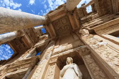 Low-angle view of the Library of Celsus façade, showing marble columns, carved stone details, and a statue against a blue sky.
