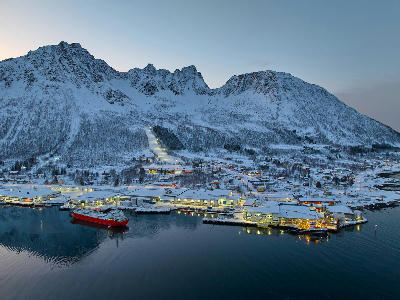 Aerial view of Senjahopen harbour at dusk with fishing boats, lit industrial buildings, and snow-covered mountains above a fjord in northern Norway