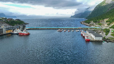 Drone view of Husøy harbour showing boats, docks, and industrial buildings with fjord and mountains beyond