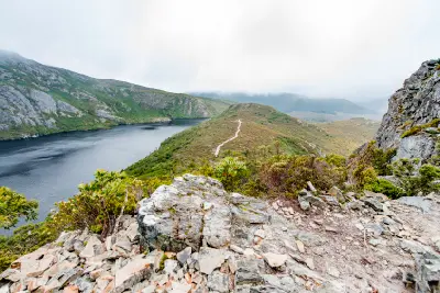 Rocky hiking trail overlooking a glacial lake and mist-covered mountains near Cradle Mountain, Tasmania.