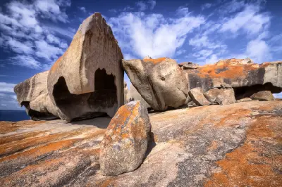 t Massive hollowed granite formations with orange lichen at Remarkable Rocks on Kangaroo Island, South Australia, beneath a dramatic blue sky with scattered white clouds.