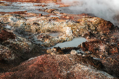 Geothermal mud pools with red and orange mineral deposits at Gunnuhver on the Reykjanes Peninsula, Iceland, with steam rising in the background.