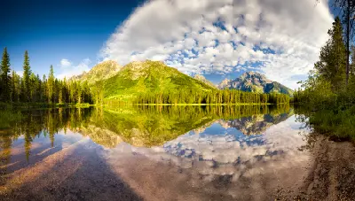Still water at String Lake reflecting green mountains, trees, and cloud-filled sky in Grand Teton National Park, Wyoming.