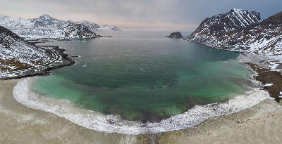 Drone view of Haukland Beach on Vestvågøya in the Lofoten Islands, showing a curved white sand bay with turquoise Arctic water and snow-covered mountains in Norway.