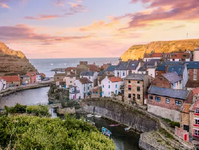 Staithes fishing village at sunset in North Yorkshire, with colourful cottages, small boats in the harbour and golden coastal cliffs beneath a pink sky.