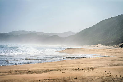 Wide sandy shoreline and breaking waves at Brenton Beach with forested hills fading into coastal mist near Knysna, South Africa.