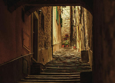 Narrow alleyway in Montepulciano, with worn stone steps, rustic buildings, green shutters, and warm light on old stone walls.