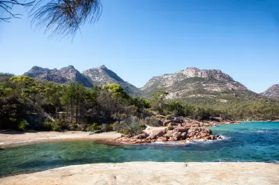 Clear blue-green water and pink granite shoreline at Honeymoon Bay with the rugged peaks of The Hazards rising behind, Freycinet National Park, Tasmania.
