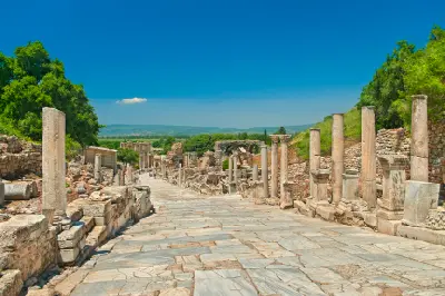Wide marble street lined with ancient columns and ruins, descending toward the Library of Celsus at Ephesus beneath a clear blue sky.