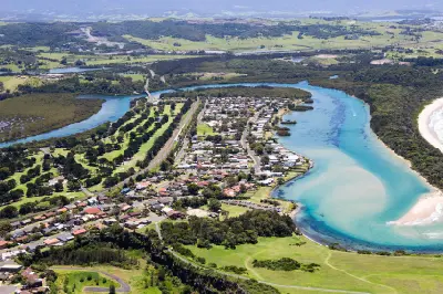 Aerial view across Minnamurra township with the turquoise river winding through wetlands toward the ocean, backed by green countryside and distant hills.