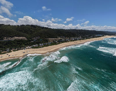 Drone view of Wilderness Beach and the Garden Route coastline with surf, sandy shoreline and green hills in the background.