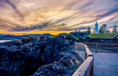 Kiama Lighthouse rises above the rugged volcanic rocks of Blowhole Point beneath dramatic streaked clouds and warm evening light.