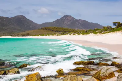Turquoise water and waves along the white-sand beach of Wineglass Bay with rugged forested mountains rising behind in Freycinet National Park, Tasmania.