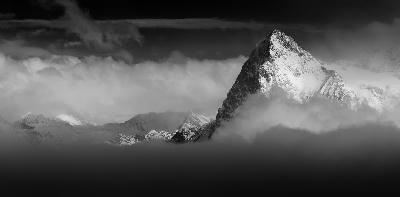 The Eiger north face rising above clouds in the Bernese Alps, Switzerland, with surrounding mountain peaks partially obscured by mist in a black and white scene.