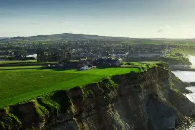 Aerial view of Whitby Abbey on a grassy clifftop above the North Sea, with Whitby town and the Yorkshire countryside in the background.