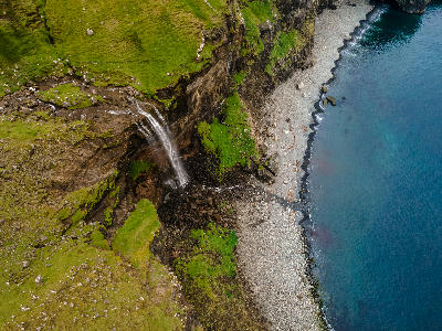 Aerial view of Skarðsáfossur waterfall flowing from green cliffs onto a rocky beach along the Atlantic coast of Vágar Island, Faroe Islands.