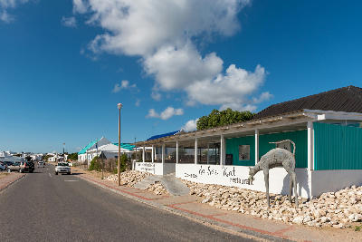 Street view of De See Kat Restaurant in Paternoster, showing a coastal village road, traditional West Coast buildings and a restaurant frontage under a blue sky.