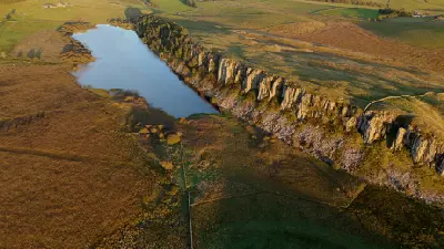 Aerial photograph of Crag Lough and the steep rocky escarpment of Highshield Crags in Northumberland, illuminated by warm evening sunlight.