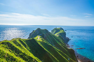 Sunlit green ridgelines and coastal path extending toward Cape Kamui with blue ocean on both sides, Shakotan Peninsula, Hokkaido, Japan.