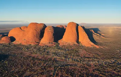 Aerial view of the sandstone domes of Kata Tjuta in evening light with Uluru visible on the distant horizon, Northern Territory, Australia.