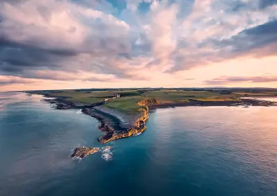 Aerial view of Dunstanburgh Castle on a rocky Northumberland headland, surrounded by the North Sea and green fields beneath dramatic pink and purple sunset clouds.