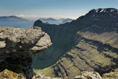 Rock ledge at Enniberg overlooking steep green cliffs and the North Atlantic, with Fugloy and Svínoy islands visible in the distance, Faroe Islands.