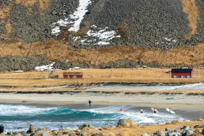 Surfers entering the water at Unstad Beach on Vestvågøya in the Lofoten Islands, with sandy shoreline, rural houses and mountain slopes behind in Nordland, Norway.