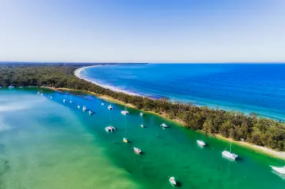 Aerial view of boats anchored in turquoise Currambene Creek beside a forested shoreline as the water opens into Jervis Bay.