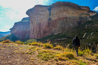 Mushroom Rock in Golden Gate Highlands National Park, South Africa, showing a massive overhanging sandstone cliff with red and cream rock layers rising above grassy slopes and a visitor standing in the foreground.