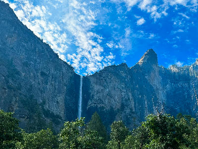 Bridalveil Fall Yosemite waterfall descending from granite cliff above green trees under blue sky with scattered clouds.