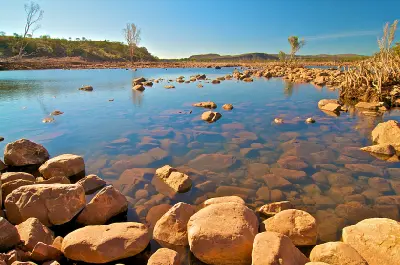 Clear shallow water flows around rounded rocks along the Pentecost River, with sparse Kimberley vegetation and distant escarpments.