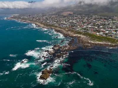 Drone view of Kleinmond on the Overberg coast showing rocky shoreline, turquoise ocean water, coastal homes, and mountains beneath low cloud.
