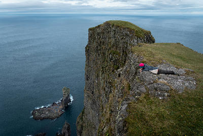 Person lying near the edge of Eiðiskollur cliff overlooking the Risin og Kellingin sea stacks in the Atlantic Ocean, Faroe Islands.