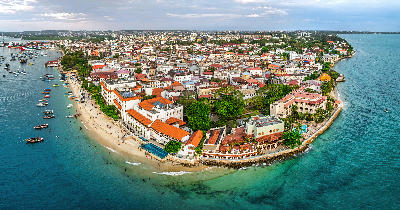 Aerial panorama of Stone Town, Zanzibar, showing historic buildings, sandy beaches, turquoise Indian Ocean waters, and traditional dhow boats.